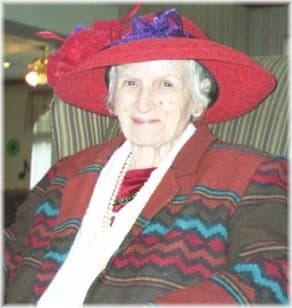 She is slim and petite with a pleasant smile and mischievous eyes. She has snowy white hair and is wearing a big red hat and a colorful jacket. She seems to be a lady with a lot of character.
