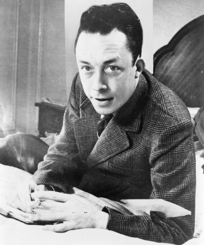 Albert Camus, Nobel prize winner, half-length portrait, seated at desk, facing left, smoking cigarette
