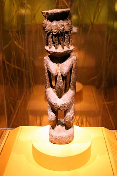 A female Dogon figure who's head is represented by an altar on which are several heads of the amphibious Nommo