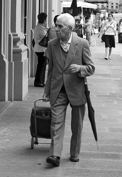 Elderly man walking with rolling suitcase and umbrella