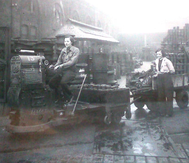 On a less than clement day, a couple of women workers pose for the camera between buildings at North Road Works, Darlington with a tractor and trailer