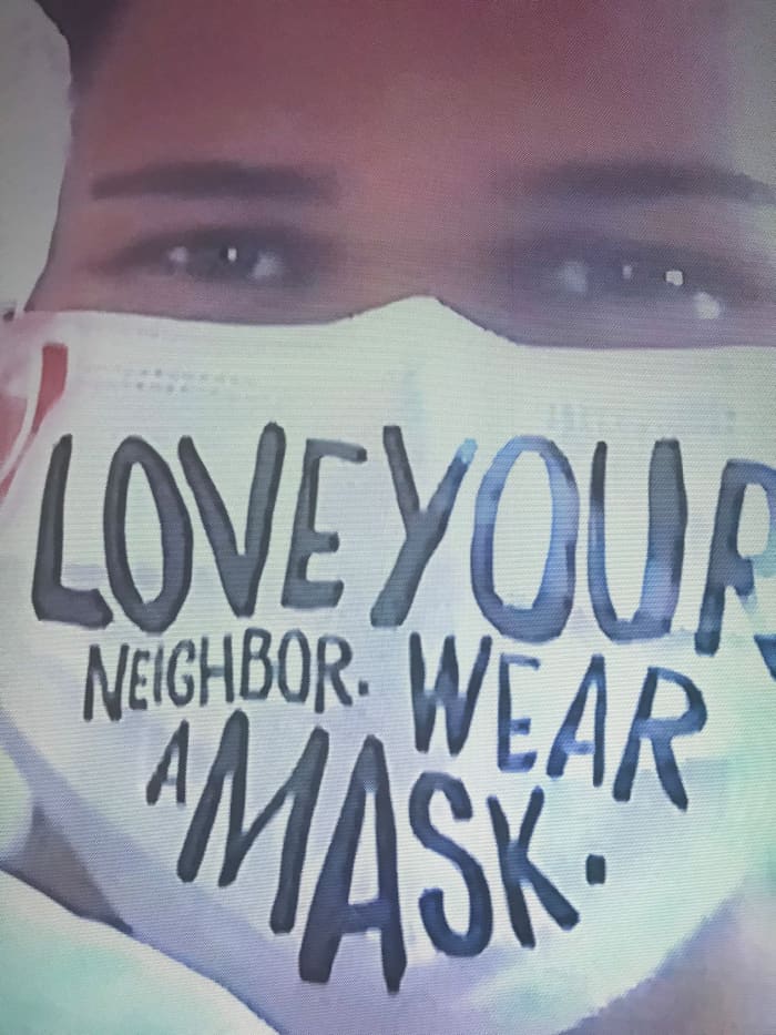 A photo of a woman from a local news story as she pleads through the message on the mask, “Love your neighbor wear a mask.”
