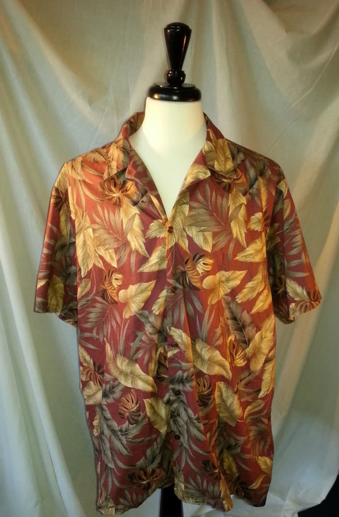 This Hilo Hattie shirt features a casual yet fun sophisticated Hawaiian print with a brick red background with a variety of gold and green leaves.