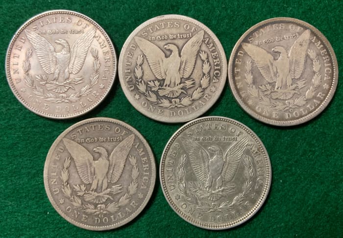 Five mint marks of the Morgan dollar below eagle. Upper-left: no mint mark for Philadelphia. Middle-top: S for San Francisco. Top-right: O for New Orleans. Bottom-left: CC for Carson City. Bottom-right: D for Denver (note how small it is).