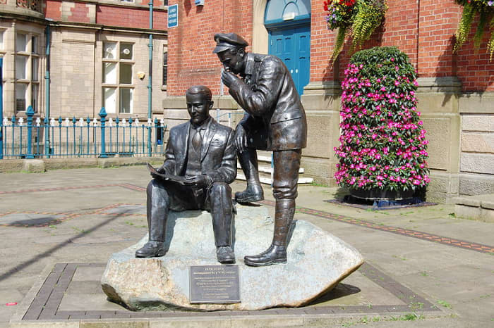 Jack Judge sits reading whilst a soldier looks over his shoulder. Statue outside Victoria Market Hall in Stalybridge.