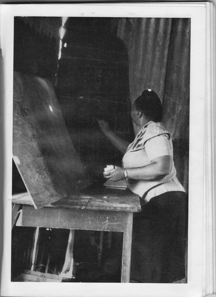 A teacher using two uneven Blackboards to write out the lesson's notes for her pupils, and the Blackboards are propped-up against the corrugated iron, and stood-up on a table. Photo #1, by Ernest L. Cole.