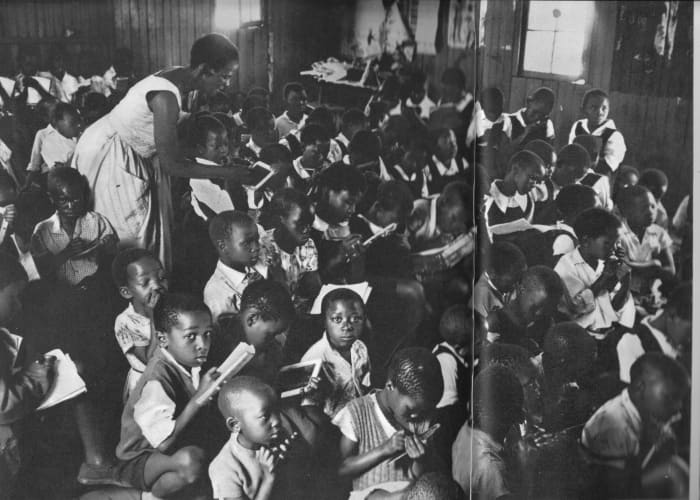 A teacher struggling with one of the two daily sessions of one hundred students each. Children learning to write hardly have elbow room to write on their slates and books
