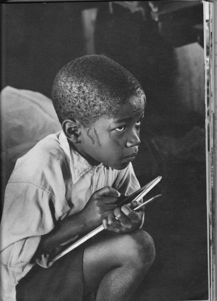 A pupil learning reading, writing and taking notes whilst earnestly squatting on haunches and straining to follow the lesson in the heat of a packed classroom