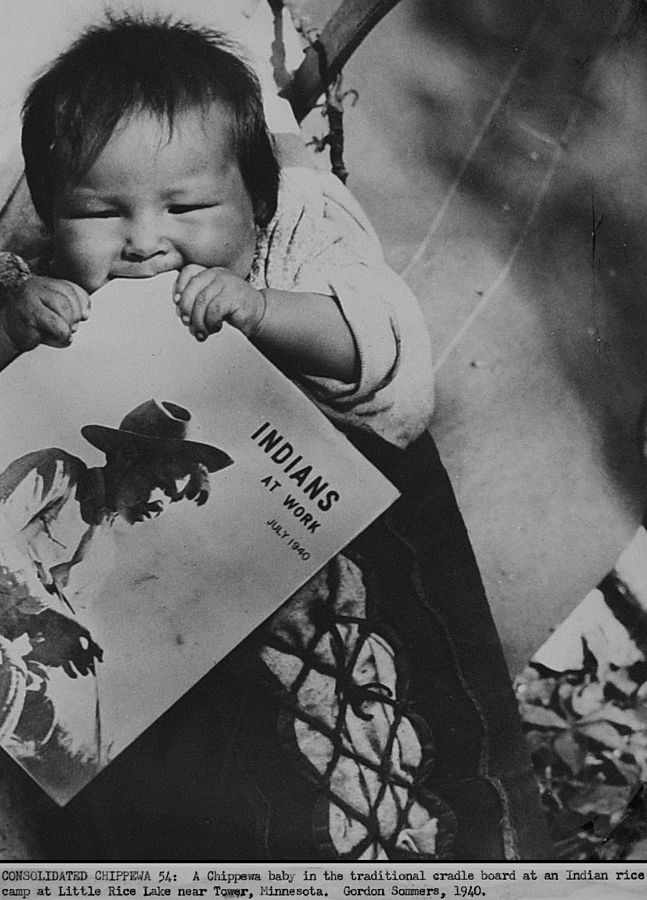 1940 US government photo. Minnesota farm worker scene of Chippewa baby (at a rice field) strapped to cradleboard while teething on an Office of Indian Affairs print magazine called "Indians at Work".