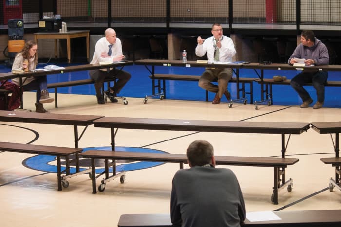 The woman to the left is showing she is timid and will be reluctant to speak up by keeping her left arm to her side and her right hand off the table, clutching the water bottle.