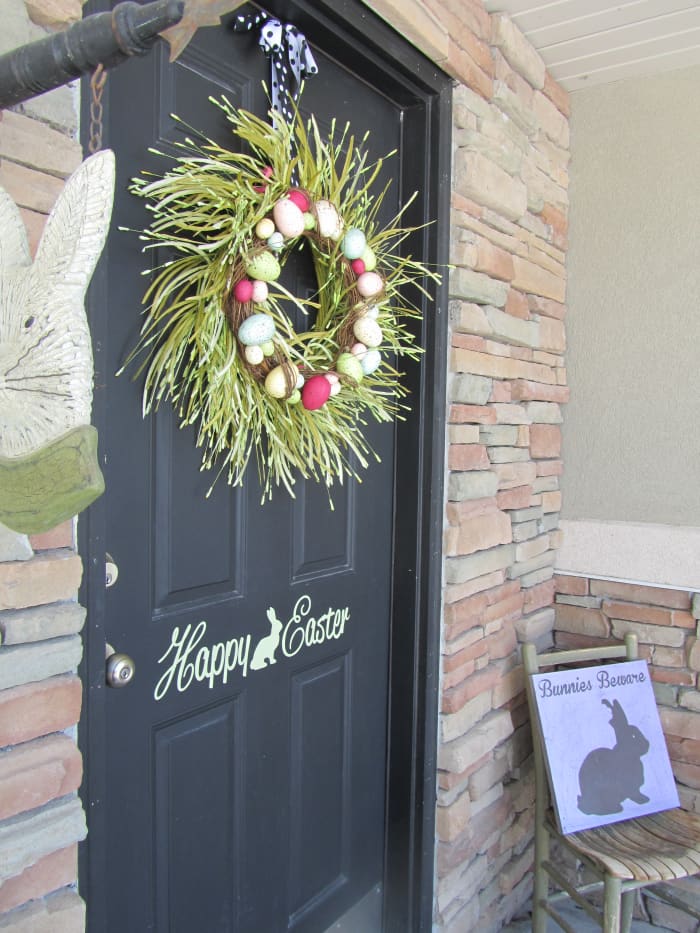 Front Door with Easter Decorations - black door with stencil Happy Easter with a bunny outline with lively green wreath with Easter eggs and Bunnies Beware sign on a small rocking chair