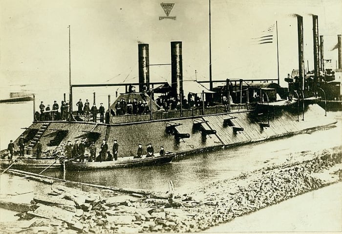 Photograph of an USS Cairo ironclad gunboat with sailors onboard. Two smaller rowboats are in the foreground with more sailors in them. In the background are other riverboats whose smoke stacks are visible.