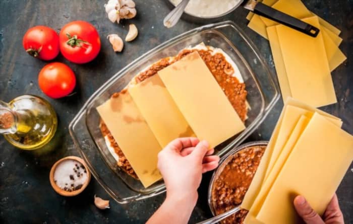 Woman cooking homemade classic lasagna bolognese, on dark blue table; with ingredients, top view copy space, hands in picture