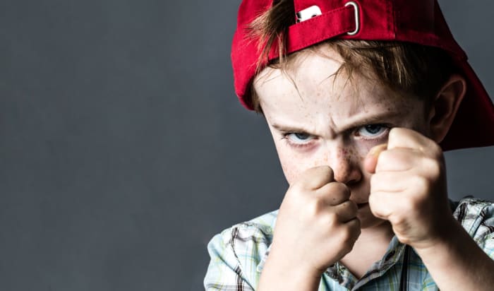 threatening 6-year old boy with freckles and a red hat back looking violent with fists in the forefront, acting like a little bully at school, contrast effects over grey background studio