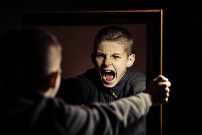 Close up Angry Young Boy Shouting on his Own Mirror Reflection with Mouth Wide Open Against Black Background.