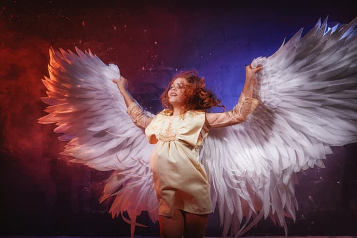 White angel on a dark background with colored lighting. The concept of war between good and evil. Girl with angel wings during a photoshoot with flour and loose powder