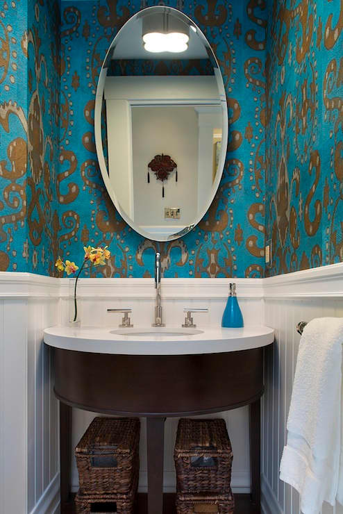 A blue powder room with a half-wall of white beadboard and ikat wallpaper in shiny gold and peacock blue. A demi-lune vanity with white counter sink with sleek faucet sits under a oval mirror in the bathroom.
