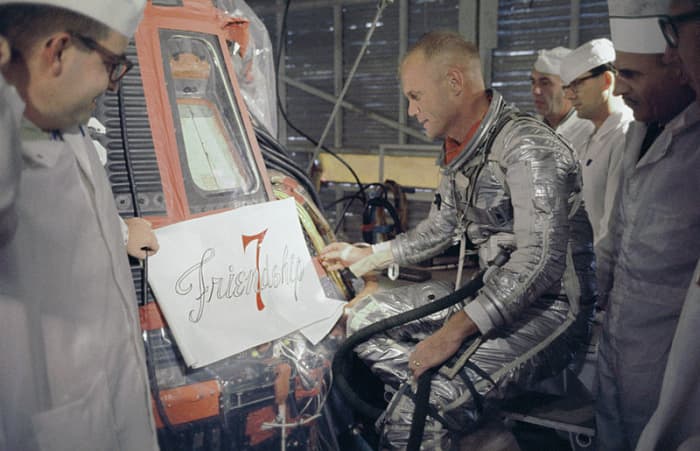 John Glenn (1921 - 2016) and NASA technicians look at the artwork to be painted on his Mercury capsule.
