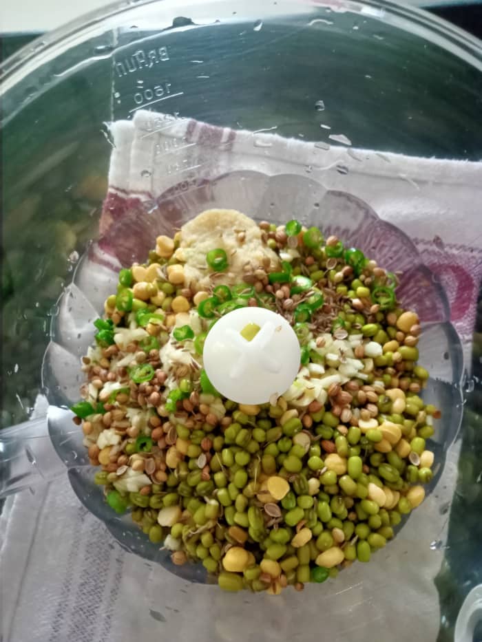 In a chopper jar, take soaked green moong and chana dal. Add chopped garlic, chopped green chillies, cumin seeds, coriander seeds, and ginger-garlic paste. Chop to a coarse paste.