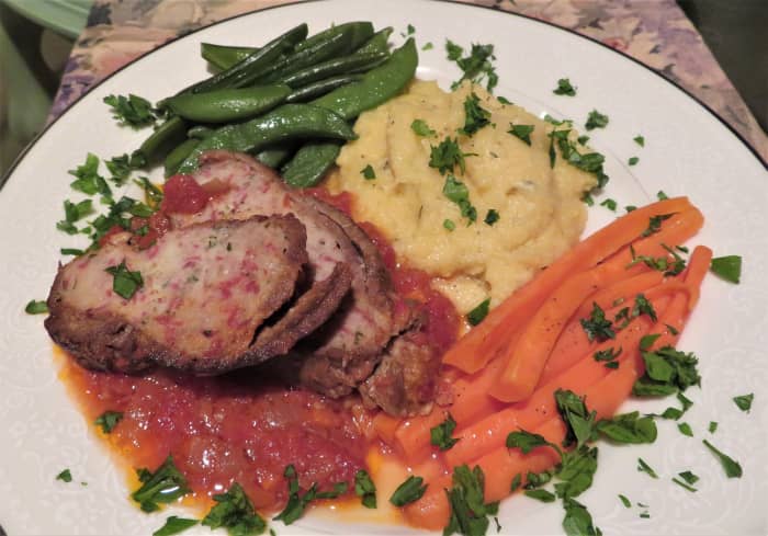 A dinner plate with two slices of braciola atop the delectable sauce, some polenta, carrots, sugar snap peas, and chopped parsley