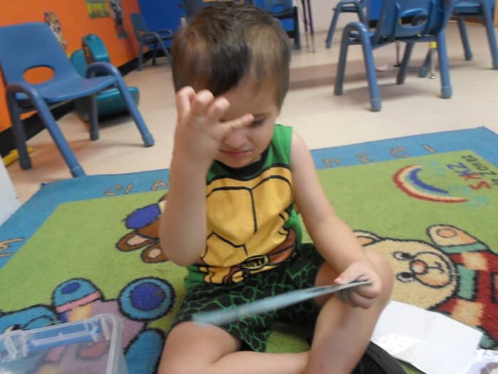 A little boy sitting on the floor with a book in his lap using sign language.