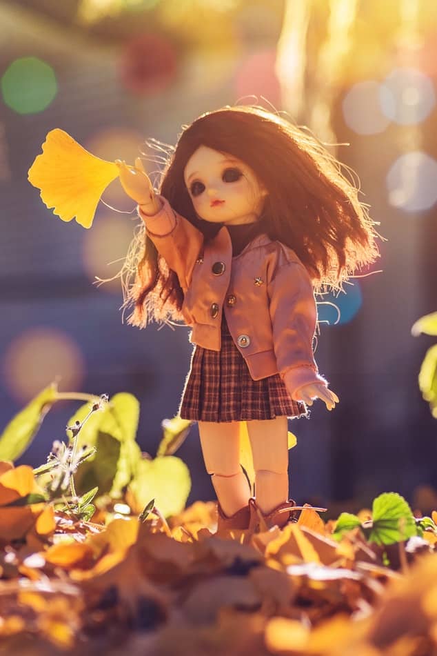 A doll standing on brown leaves is wearing an outfit of a brown skirt and jacket for the autumn weather.