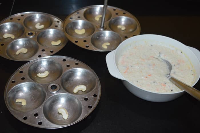 Step four: Grease the hollow containers of the idli maker with oil or ghee. Keep the steamer or cooker ready for making idlis. Place a cashew nut on each of the hollow containers. Add the batter until the brim.