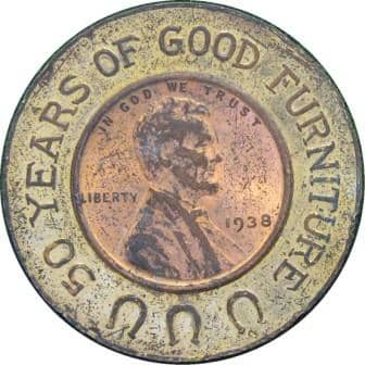 1938 Lincoln cent encased in a brass holder. The obverse features three horseshoes and the legend, “50 YEARS OF GOOD FURNITURE.”