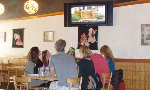 Restaurant table with patrons watching TV. Tables fill the front of the restaurant and run down the sides of both walls on either side of the sushi bar.