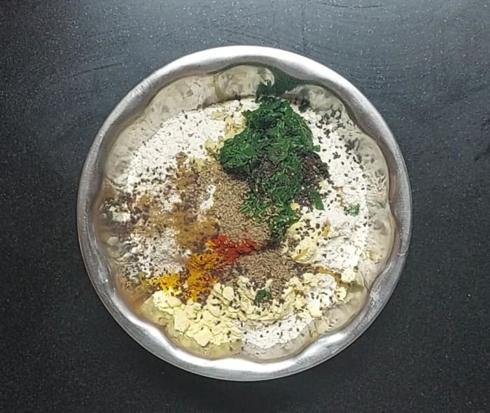 In a bowl, take 2 cups whole wheat flour, 2 tbsp gram flour, salt, 1 tsp ghee, 1 tsp grated ginger, fried methi leaves, 1/4 tsp turmeric powder, 1/2 tsp cumin powder, 1/2 tsp red chili powder, 1 tsp black sesame seeds, 1 tsp crushed ajwain.