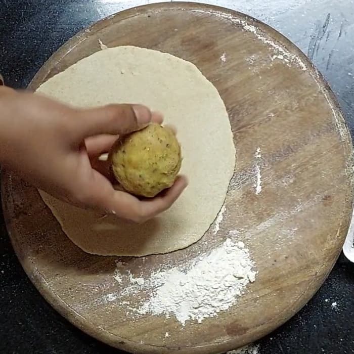 Place a dough ball on a rolling board, dust with some wheat flour and roll a small chapati using a rolling pin. Place a potato stuffing ball in the center.