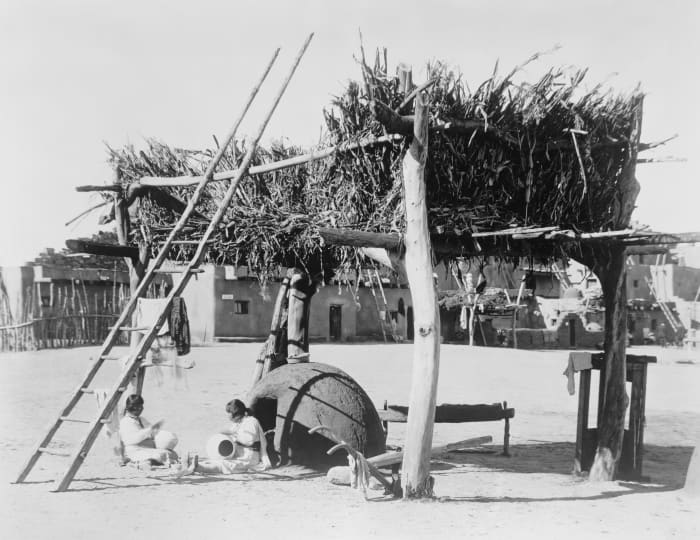 1915 photo of Zuni women crafting pottery under a drying rack for maize and other foods. Note the horno (outdoor adobe oven) which serves a dual purpose: drying the plants and to glaze the pottery.