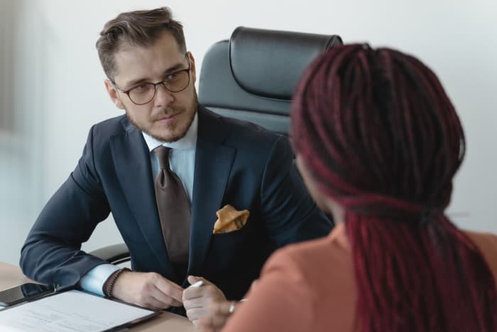 A man in a black suit sits in an office chair and looks over a woman opposite his table. The final interview can be scary, exciting, and useful all at the same time.