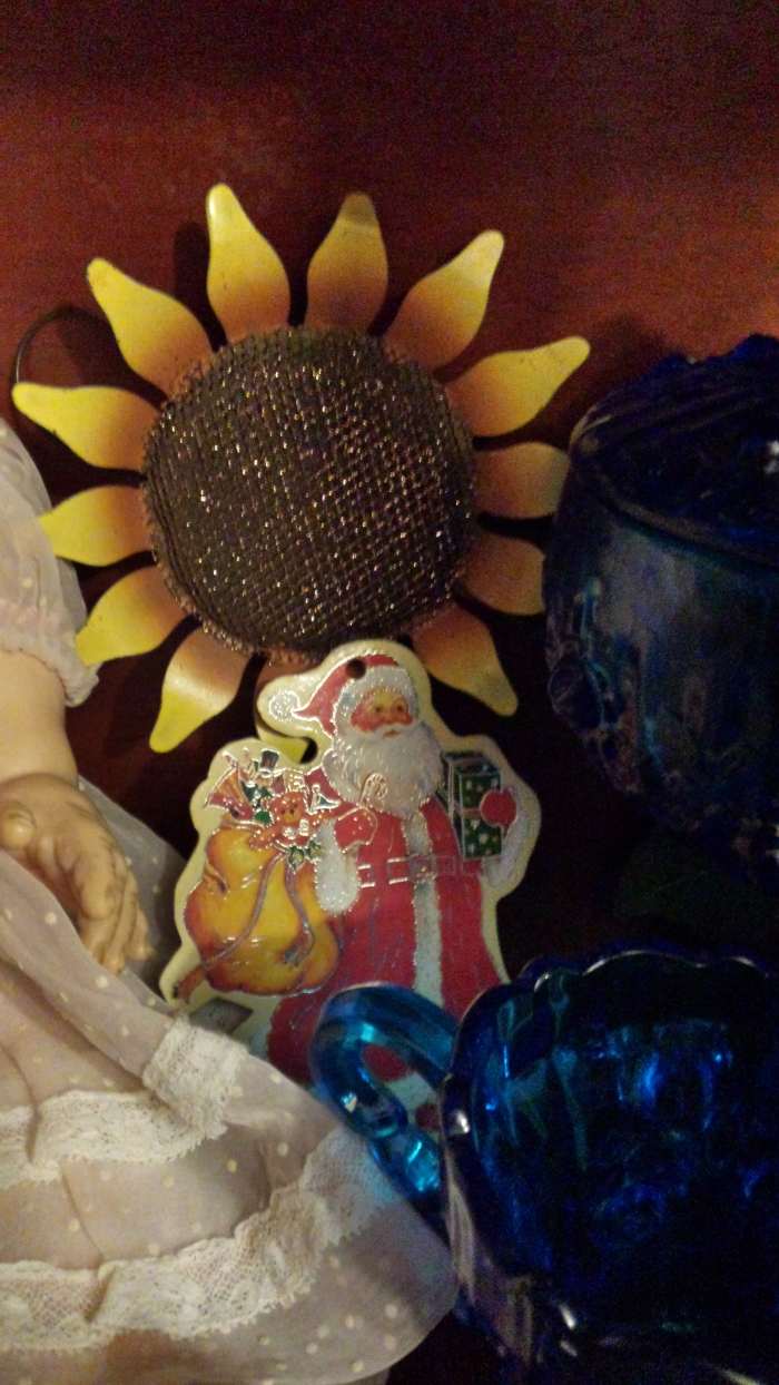 Photograph of vintage cardboard cutout Santa ornament with gold outlines. He stands in front of a metal sunflower and beside antique blue glass. There is a baby doll seen only in part on the photo's left side.