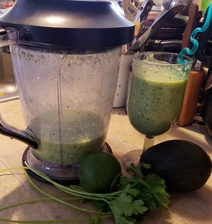 A glass full of this refreshing green smoothie, accompanied by some of the ingredients and the Ninja used to make it.