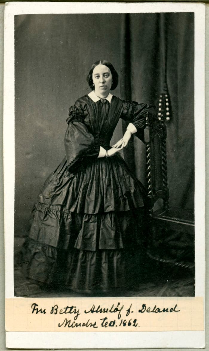 1860s: Pagoda sleeves, low set shoulder line, flounced hoop skirt, and central part in her hair suggest this picture was taken in the 1860s.