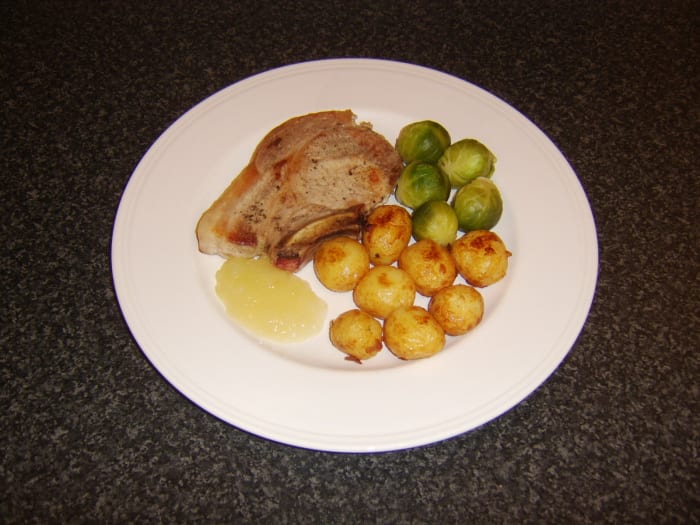 Pork chop shallow fried in oil and served with roasted baby potatoes, Brussels sprouts and apple sauce