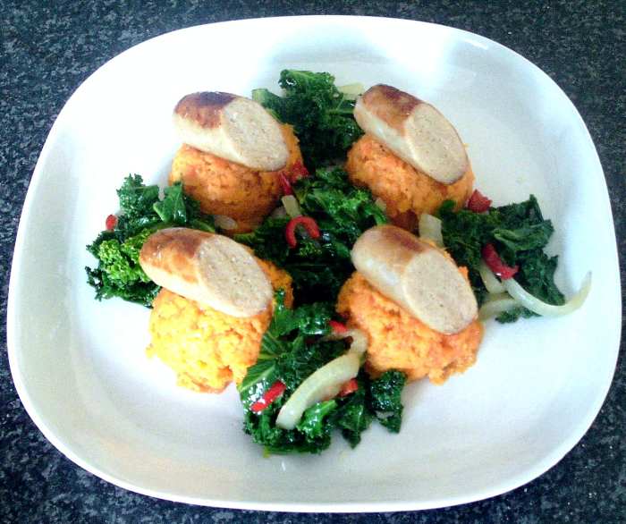Chicken sausage halves are arranged on scoops of sweet potato and carrot mash, served with chilli and garlic spiced sauteed kale