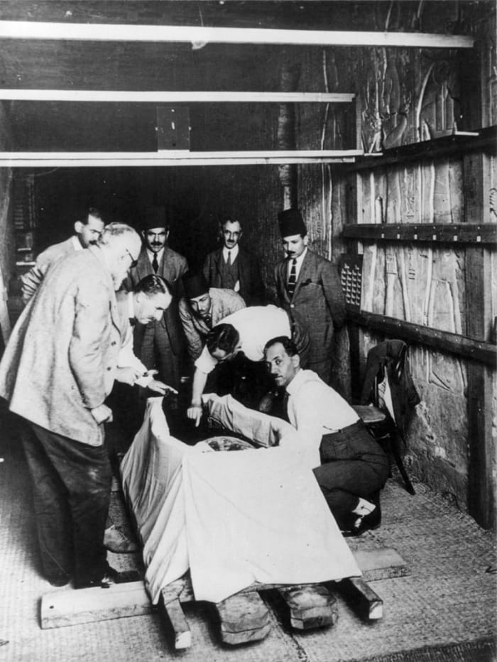 Howard Carter, with magnifying glass, leaning over Tutankhamun during the unwrapping of his mummy in 1925.
