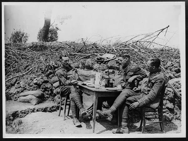 British officers mimic "fine dining" in reserve. There are flowers, mugs, plates, and a bottle labelled "Dark Port" on the table but no food can be seen.