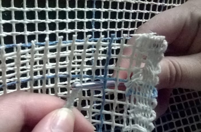 Using a crochet hook or other instrument, pull one of the long strands of yarn from your pom pom through one of the squares in your canvas. Notice how the intersection of the blue lines forms an X shape.
