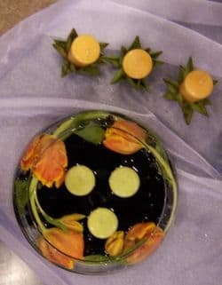 Orange parrot tulip and artichoke reception centerpiece with black stones and green candles in a low glass bowl.
