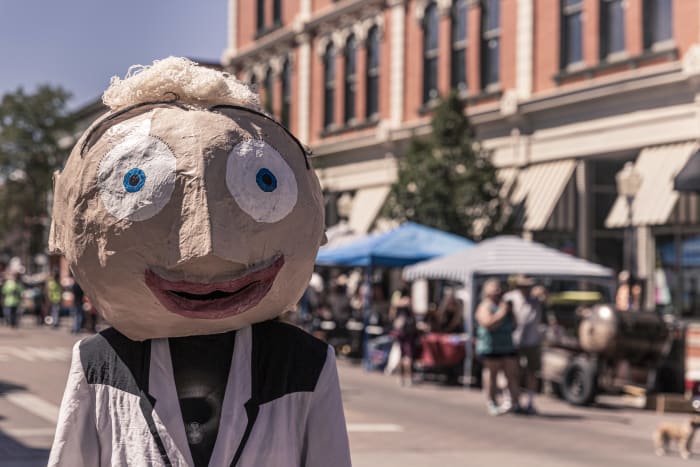 A young man wearing an over-sized paper mache head as a mask to complement a wrinkled lightweight jacket and tee shirt.