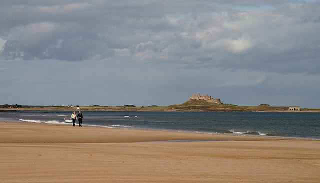A couple walk along the shoreline with Lindisfarne Castle on Holy Island in the background.
