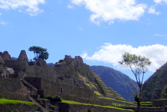 Terraced ruins being explored by a few tourists, with trees framing the brilliant sky.
