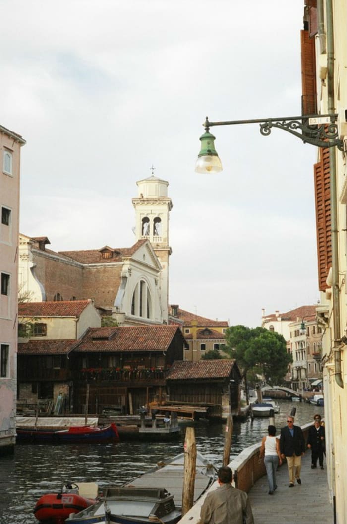 Pedestrians walk alongside the canal near the Squero di San Trovaso boatyard, where gondolas are made, with the Church of San Trovaso in the background.