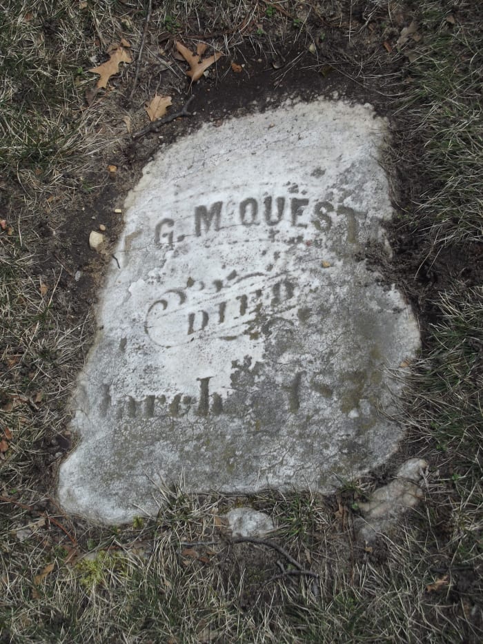 A somewhat square tombstone flat on the ground that is mostly intact. The text is relatively faded. The grass has grown over the sides. Evergreen Cemetery, Muskegon, MI.