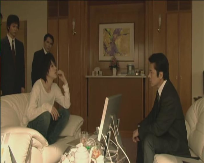 L's clothing, posture and mannerisms contrast with the officers in full suits and natural postures.