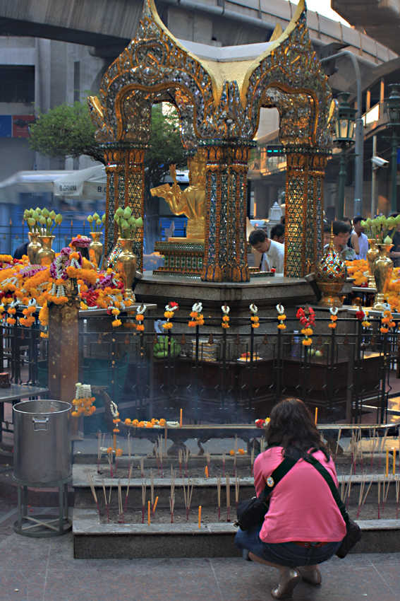 A girl prays in front of the shrine. The smoke from the incense is clearly visible as a blueish haze. Top of the picture is the concrete support of the overhead skytrain - the sounds of the metropolis are so close