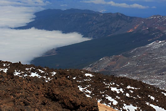 The view northeast from the upper station. Note the red lava flows in the foreground and the dark lava flow in the centre, the low cloud bank, and the 'Cordillera' Mountain Range in the distance